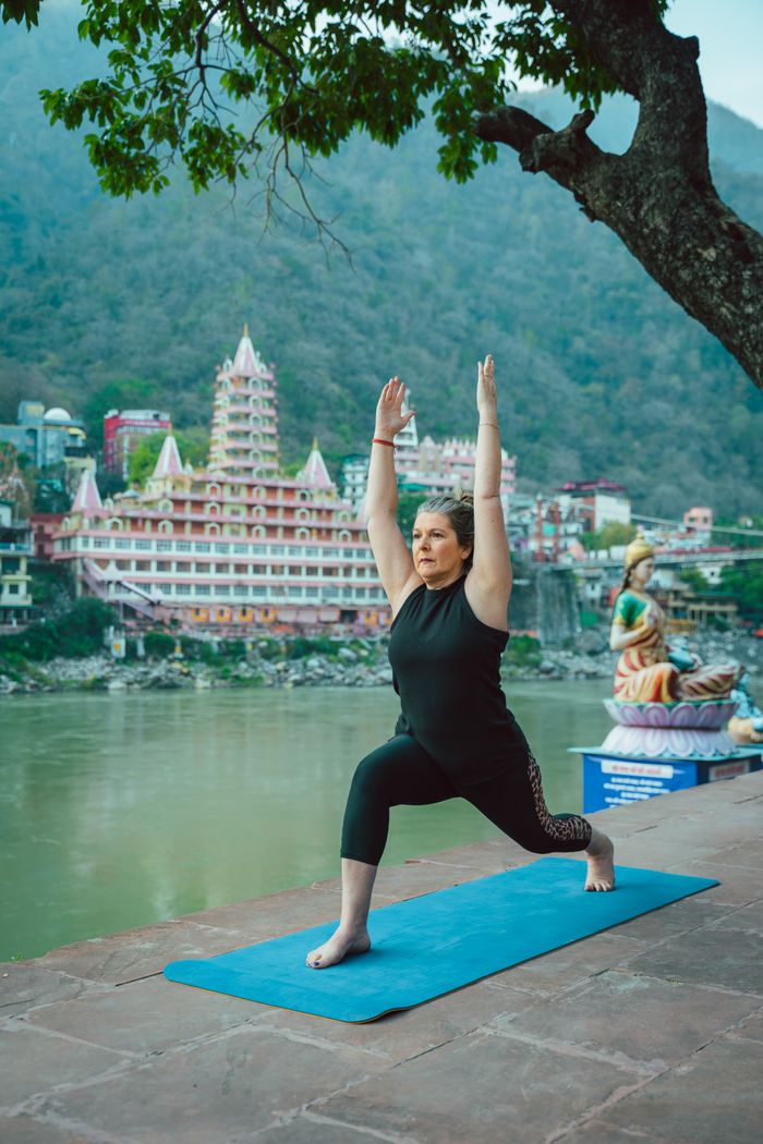 A woman doing yoga by the Ganges river with a scenic view of Rishikesh, India.