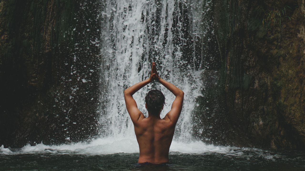 Offerings A man stands in a meditative pose by a waterfall, showcasing tranquility and nature adventure.